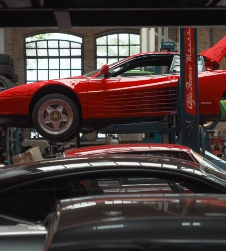 About A vibrant red sports car is elevated on a lift inside an auto repair garage, surrounded by various vehicles and equipment.